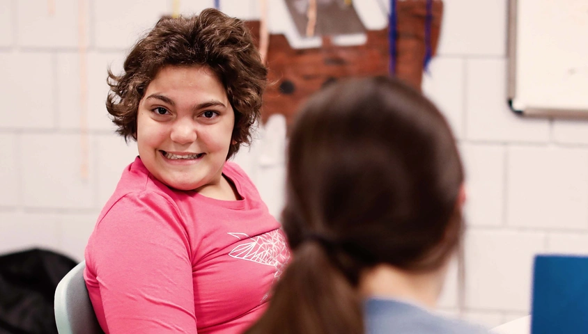 Caregiver in foreground facing away from camera, client in background smiling at camera. 