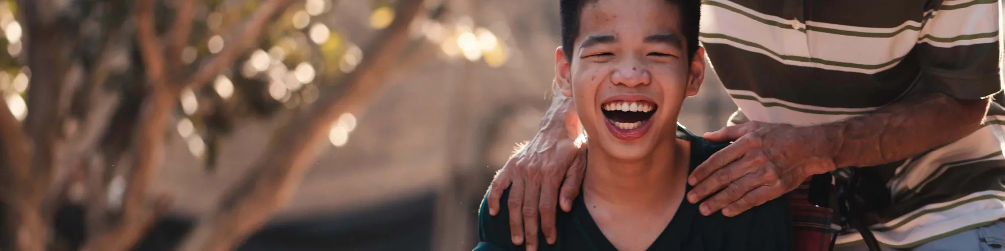 Child outside in wheelchair, laughing, adult behind holding wheelchair laugh while looking at child. 