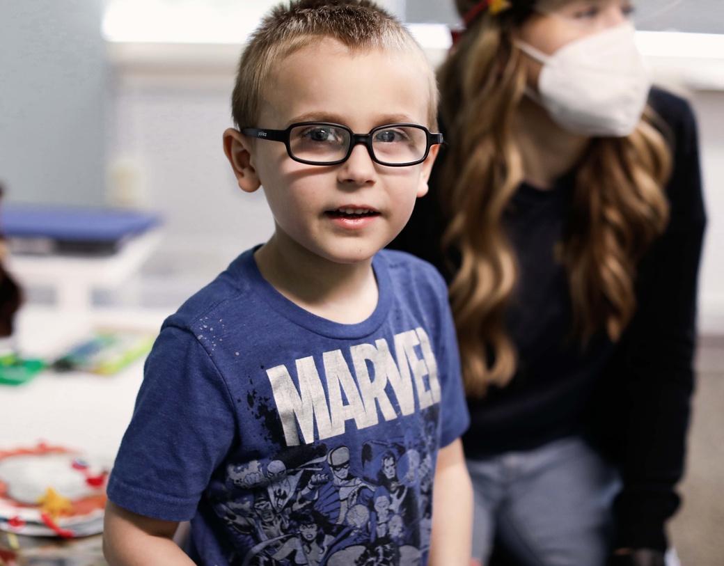 Child wearing glasses looking at camera, parent in the background with art supplies. 