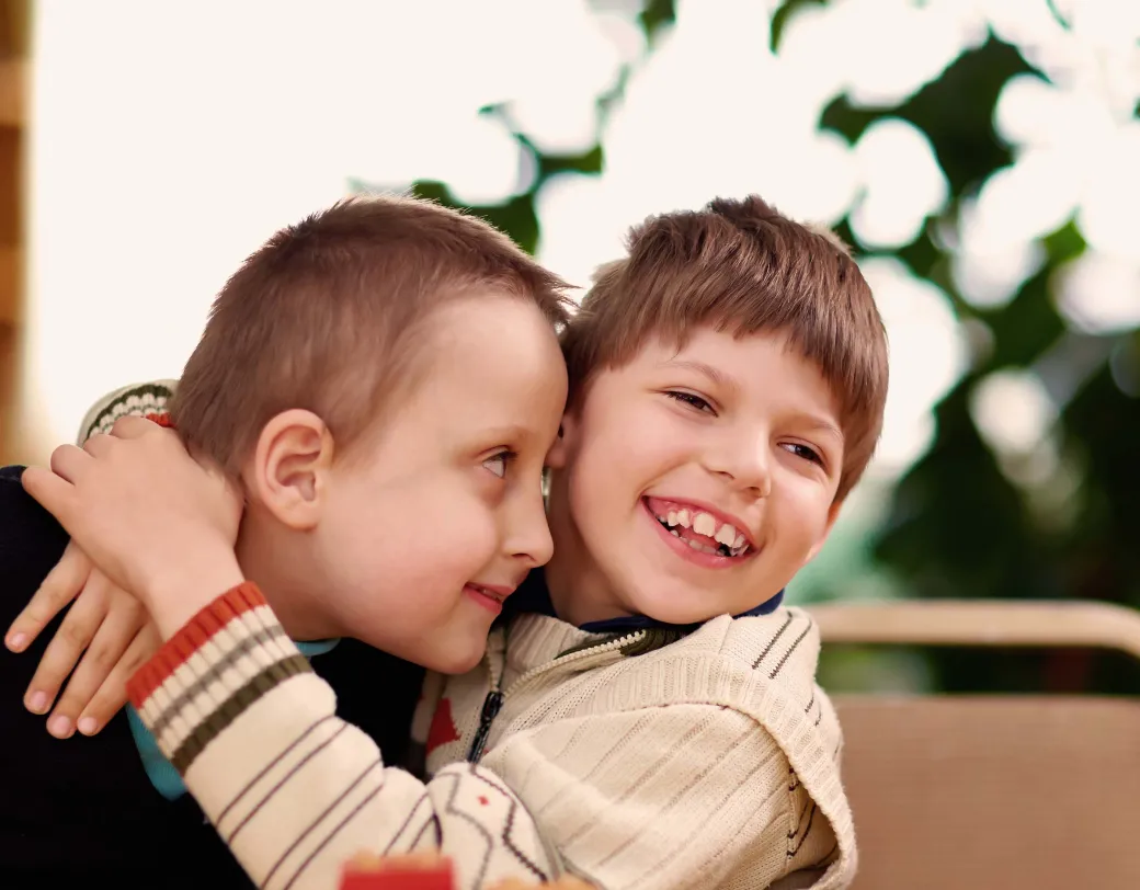 Two children, one with arm around the other, sitting at table with colorful block toys. 