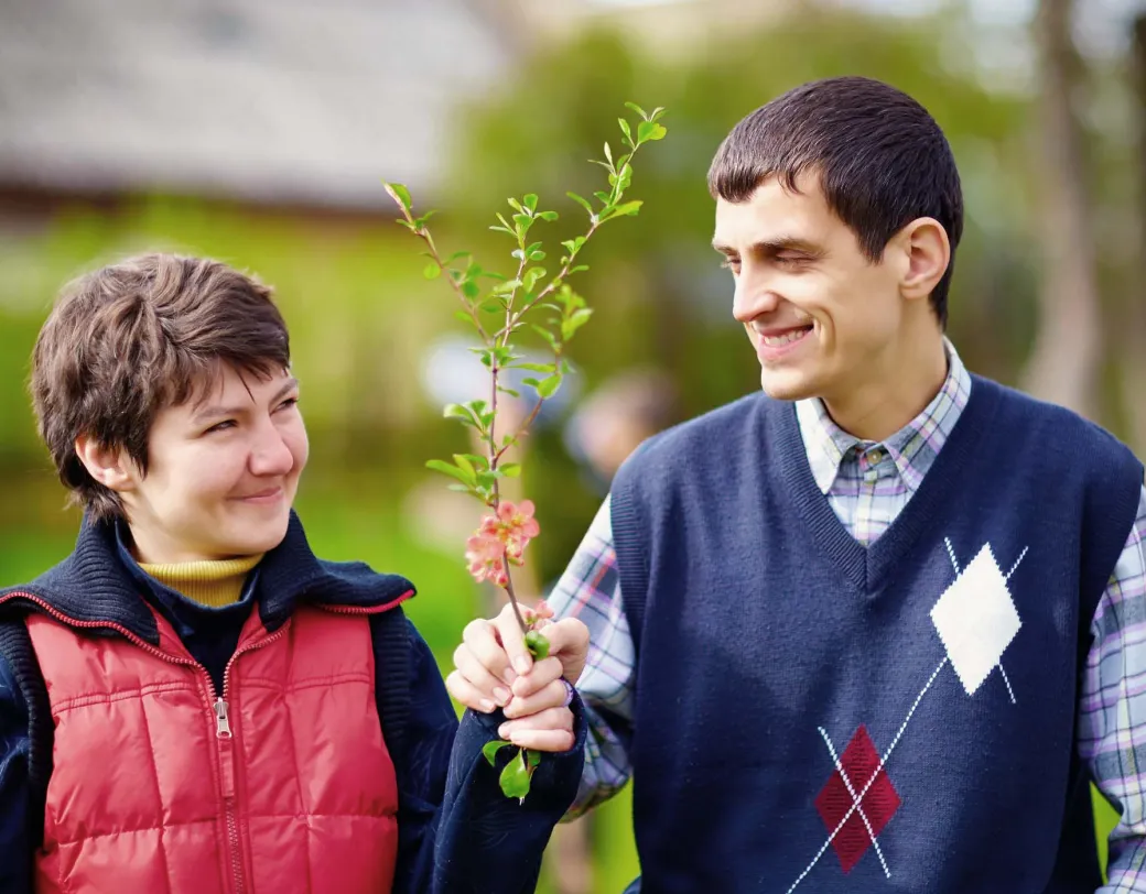 Two adults walking together outside. One person handing the other a branch with leaves and flowers.