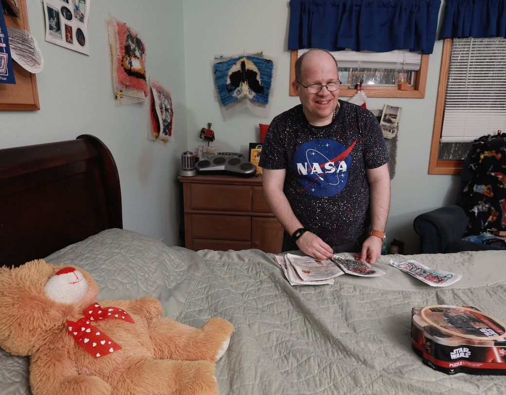 Person standing next to bed, in bedroom, holding artwork and smiling.