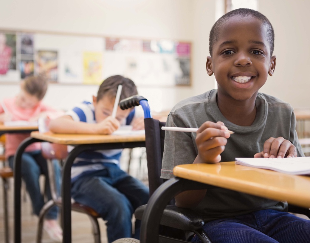 Child sitting at desk, smiling, other children at desks in background. 