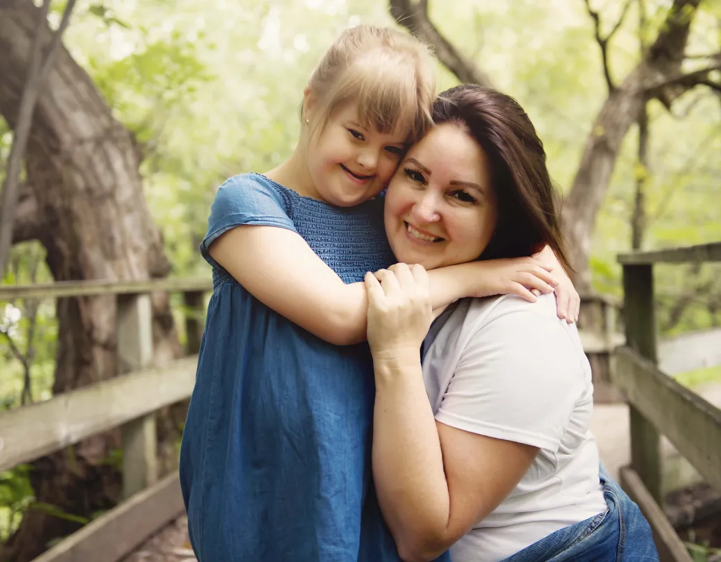 Parent and child hugging on wooden bridge outdoors in wooded area. 