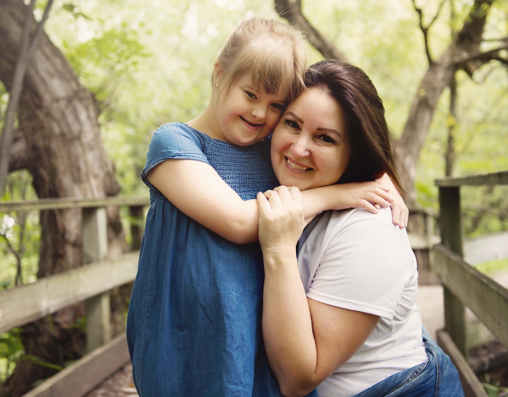 Parent and child hugging on wooden bridge outdoors in wooded area. 
