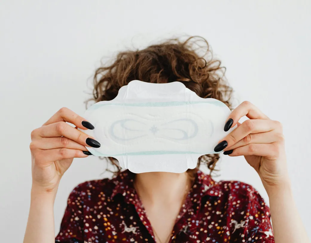 White woman with curly brown hair holds unused sanitary pad open in front of her face.
