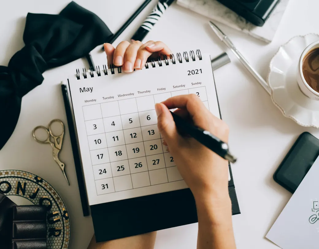Person writing on a monthly calendar with a black pen. Things are scattered in the background.