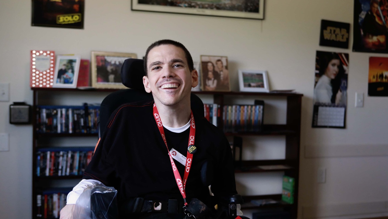 Person smiling in wheelchair in their living room, wearing work uniform.