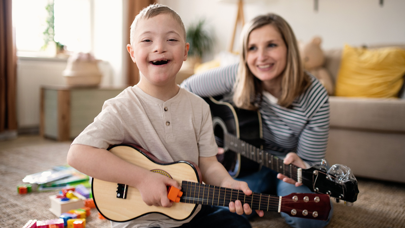 Child and adult care provider sitting on living room floor playing acoustic guitars.