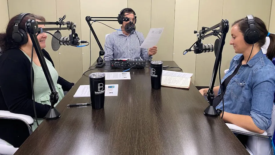 Scott Light at the head of a podcasting table. Andrea Ryan and Alicia Boreman on either side of the table speaking into microphones. 