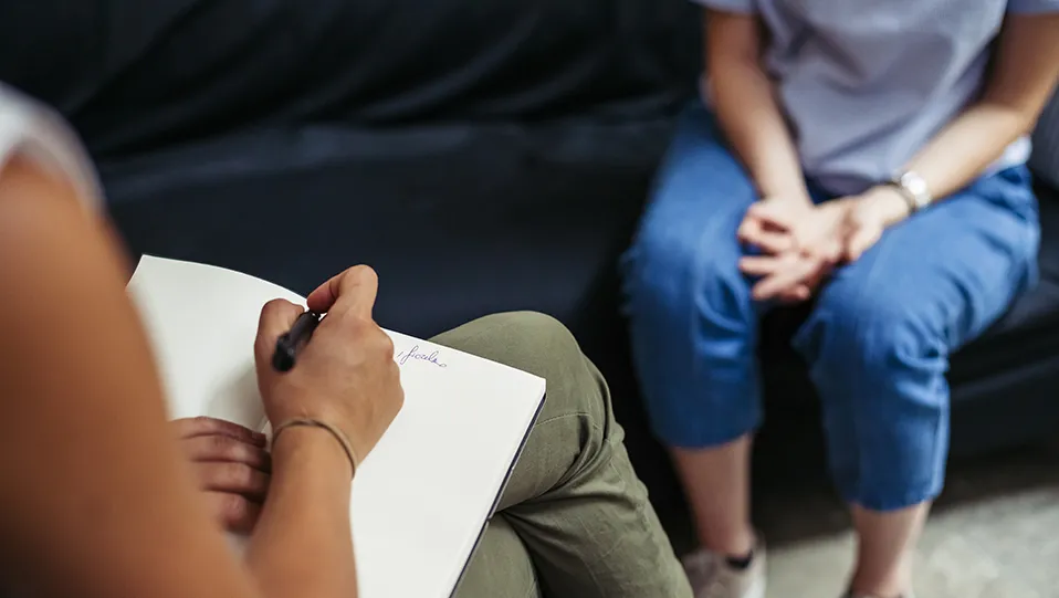A person on a couch with folded hands across from a counselor taking notes.