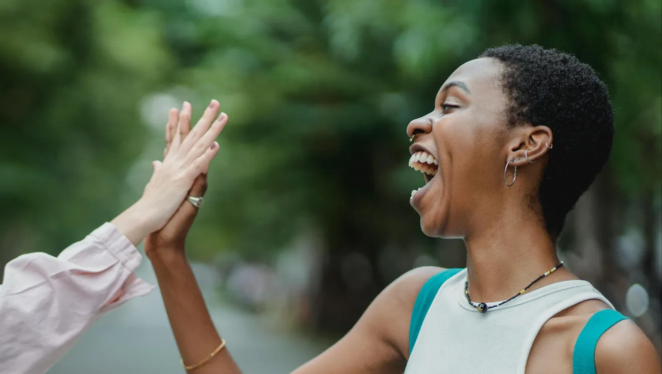 One black woman high fives another person who is white and out of frame except for their arm. They are outside.